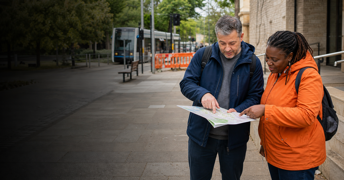 A visitor and a community access coordinator compare two routes on a town map near a tram stop, bench, temporary barrier, and level entrance